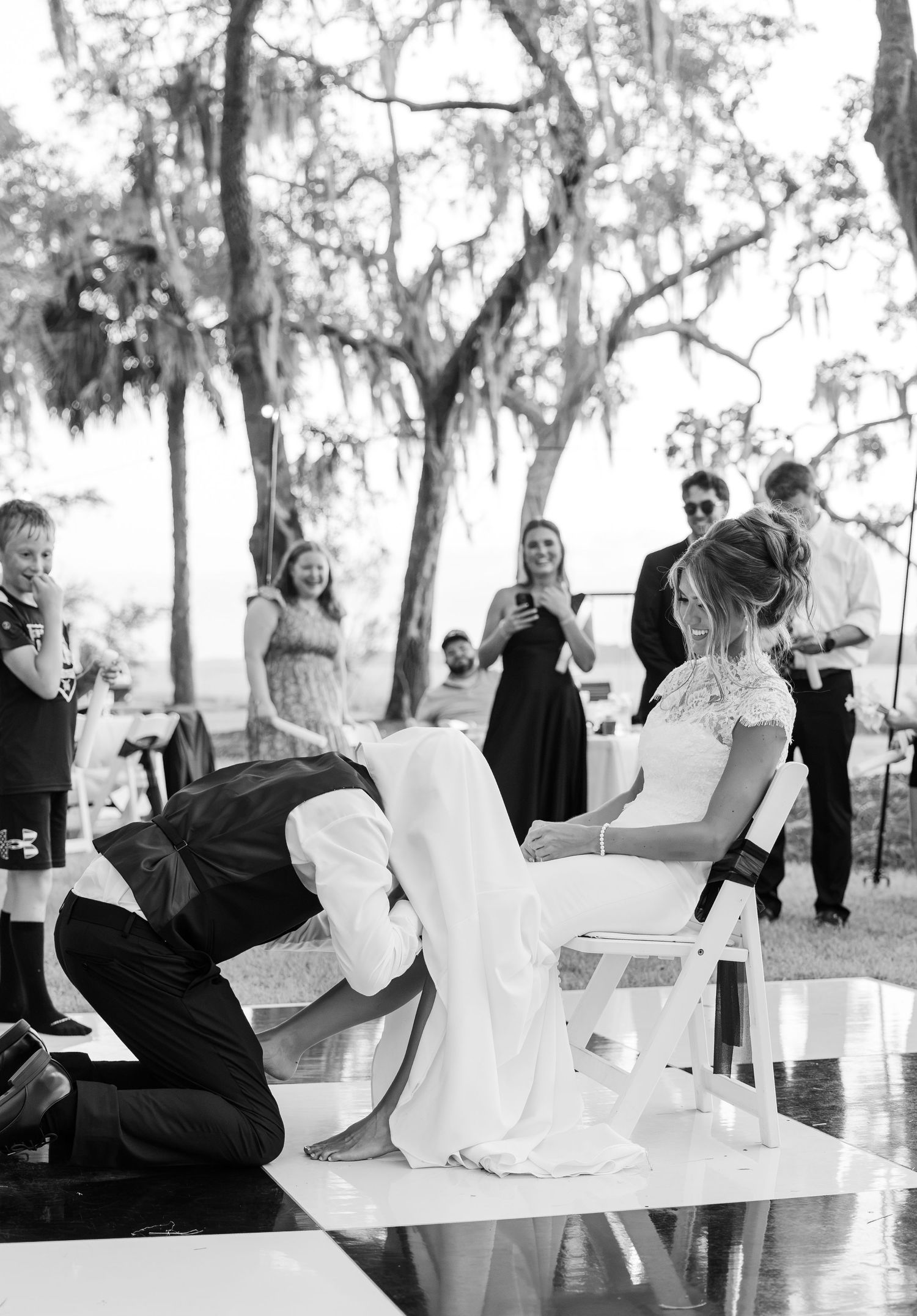 Groom removes bride's garter during outdoor wedding reception while guests watch and smile.