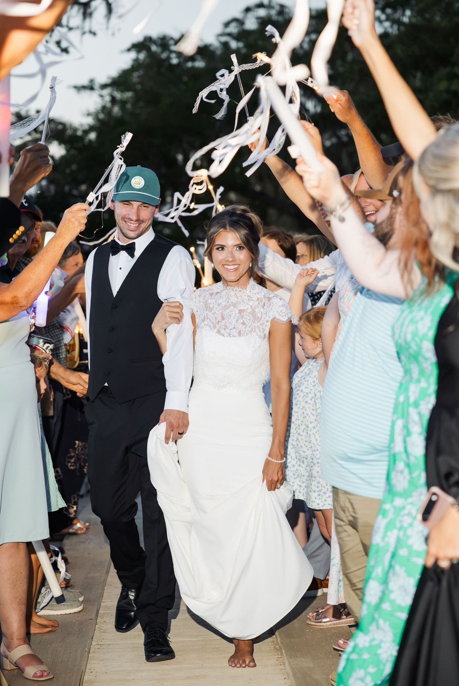 Bride and groom walk through ribbon send-off tunnel surrounded by cheering wedding guests at outdoor evening reception.