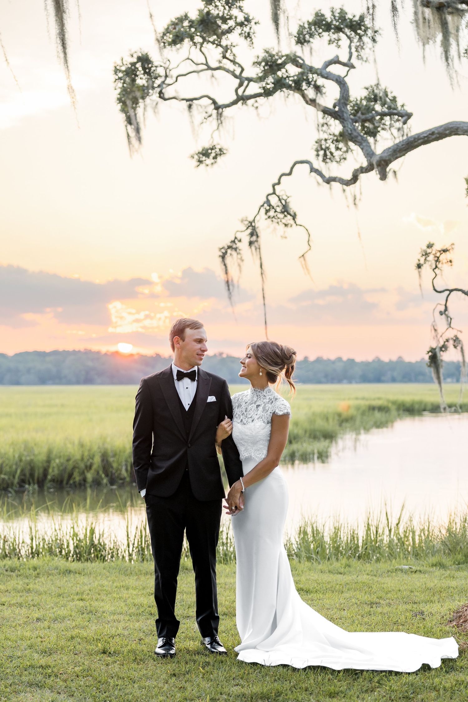 Bride and groom hold hands at sunset by a marsh, framed by Spanish moss-draped oak trees in a romantic outdoor wedding portrait.
