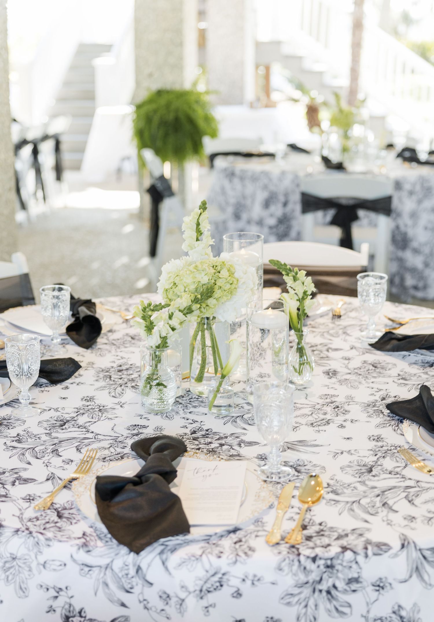 Elegant wedding reception table with black and white floral tablecloth, gold cutlery, black napkins, and white floral centerpieces.
