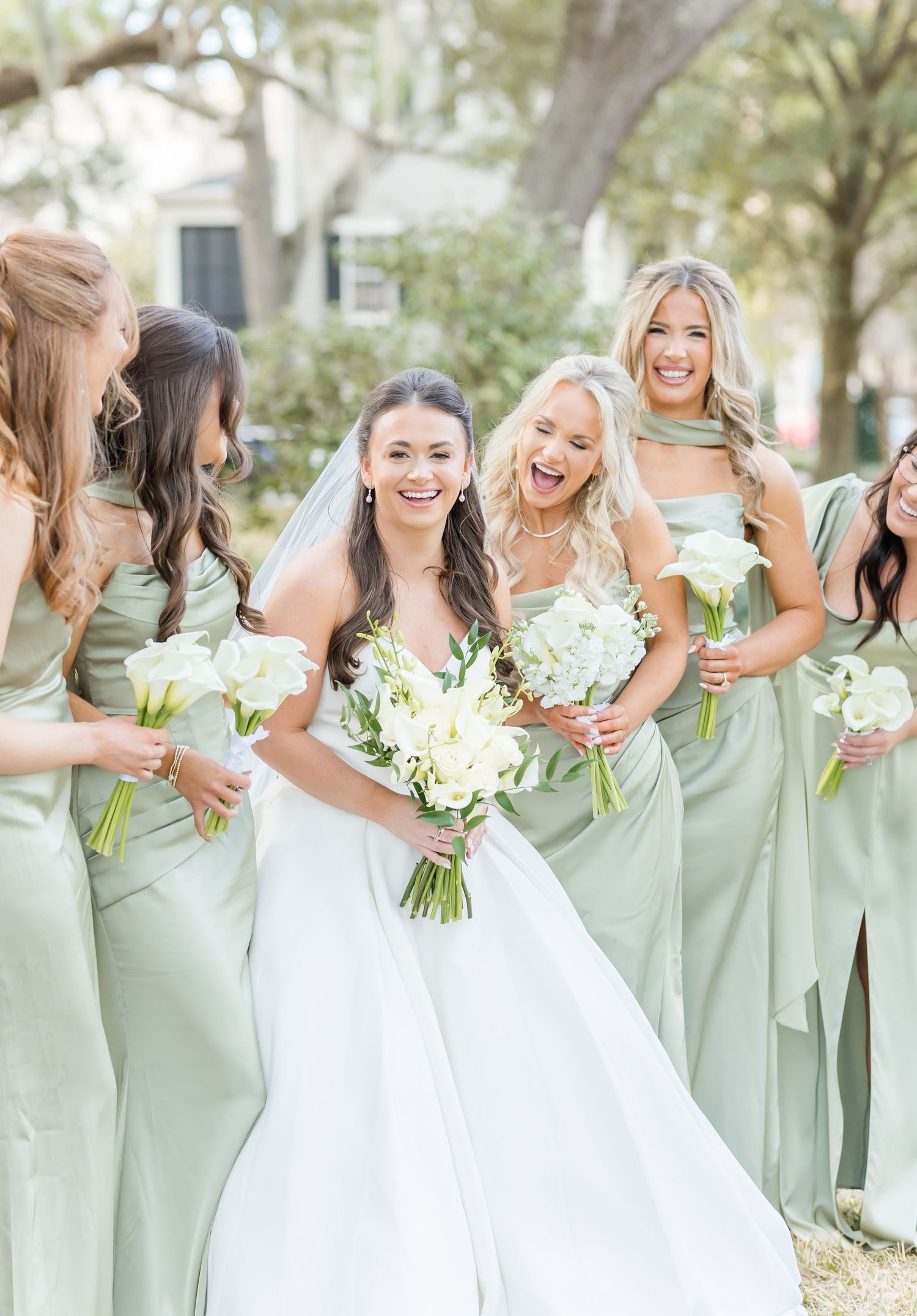 Bridesmaids in soft sage green dresses share candid laughing moments with the bride in a white gown outdoors.
