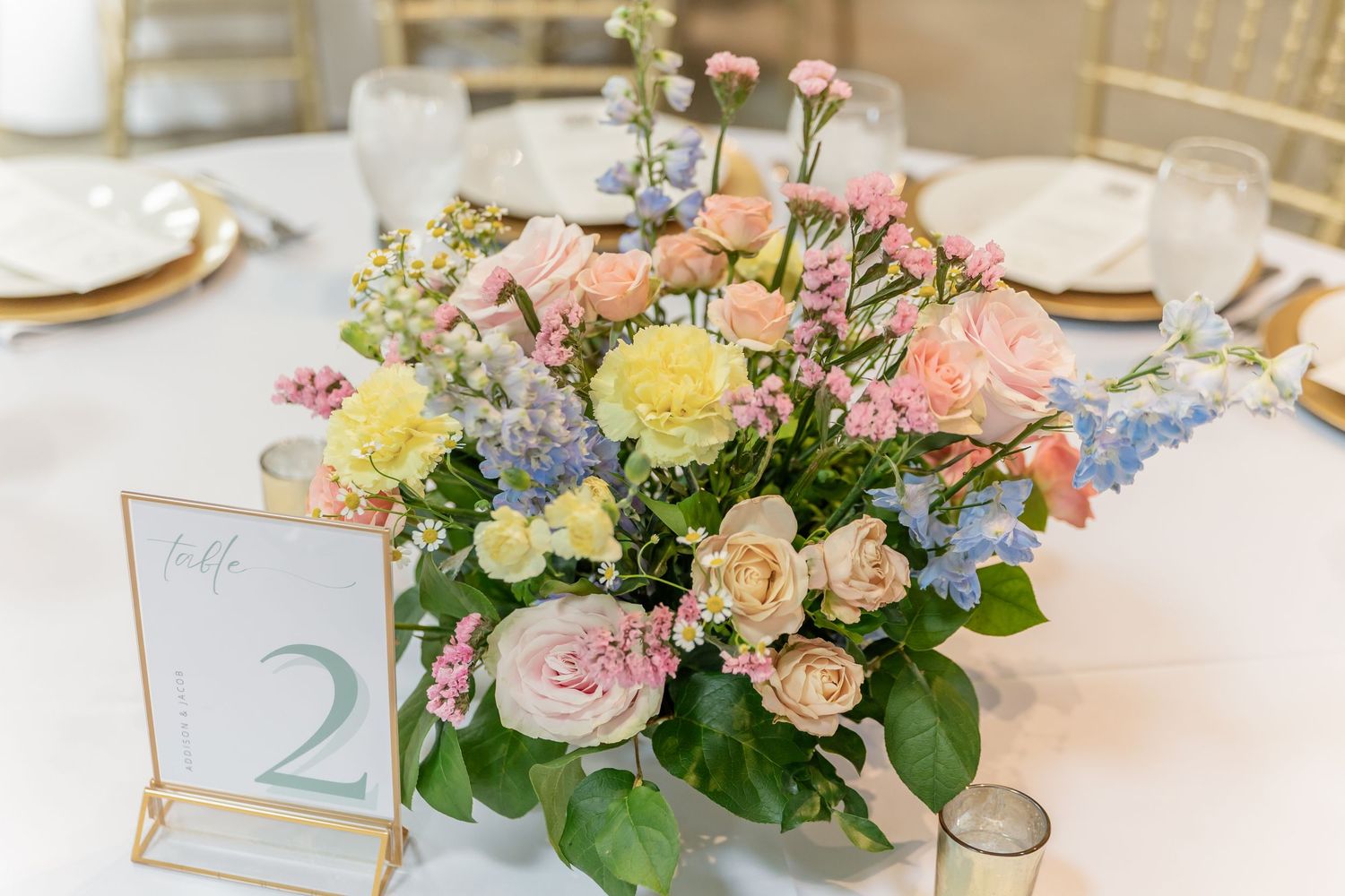 A pastel floral centerpiece with roses, yellow blooms and blue flowers on a white tablecloth with a table number card.
