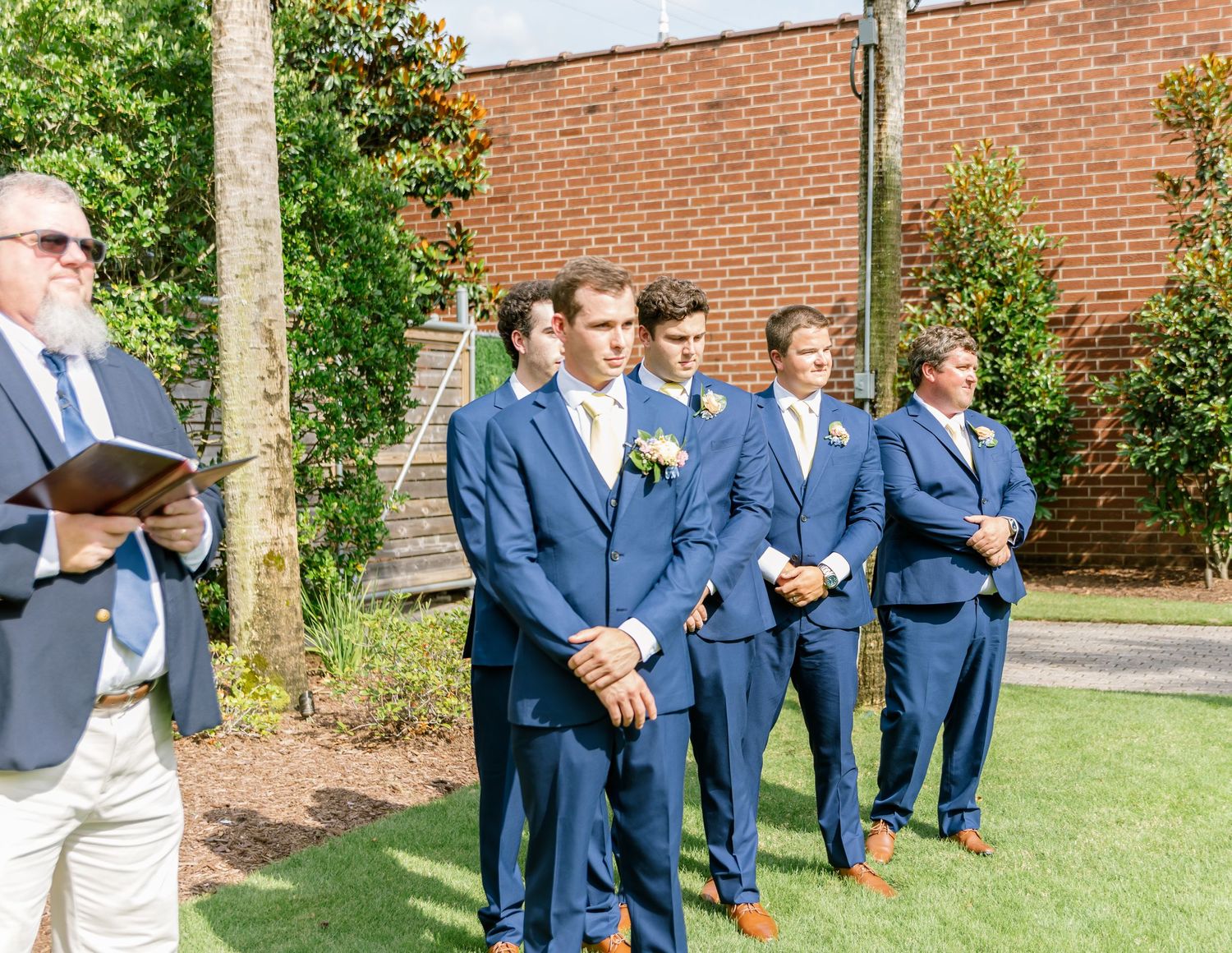 A line of groomsmen in matching blue suits stand outdoors by a brick wall during a wedding ceremony.