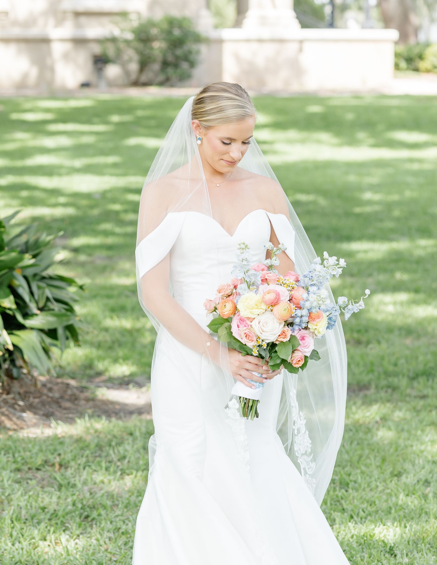 A bride in a white wedding dress holds a colorful bouquet of pink and peach flowers while standing in a sunny garden.