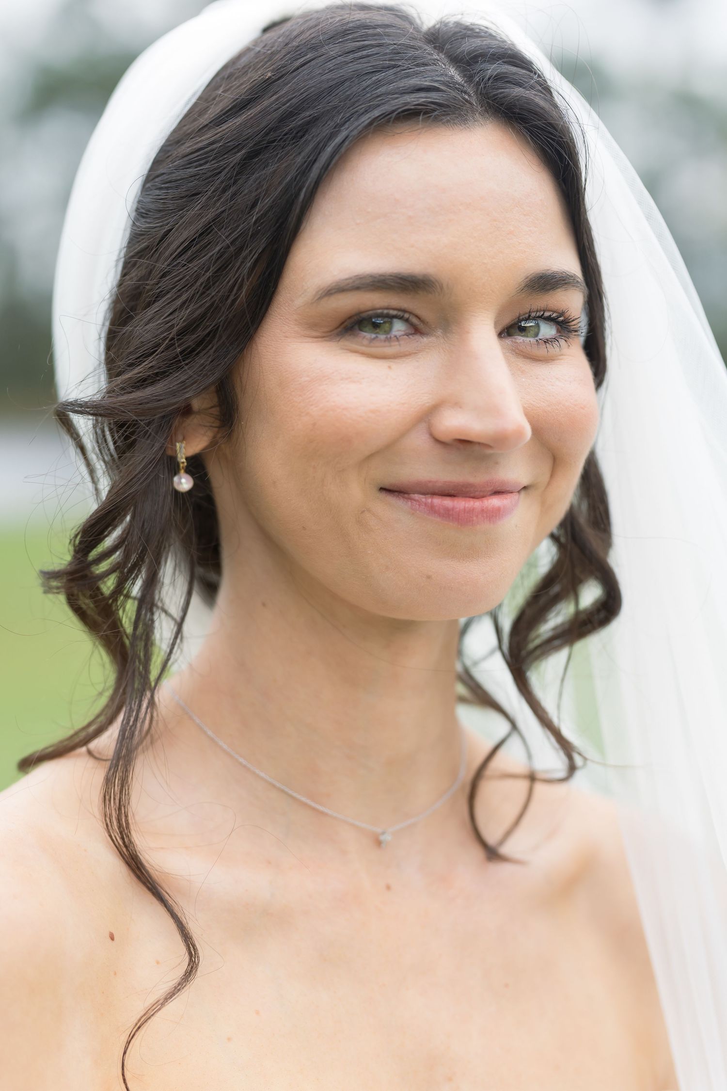 A bride wearing a white veil and delicate jewelry smiles softly in an outdoor portrait.