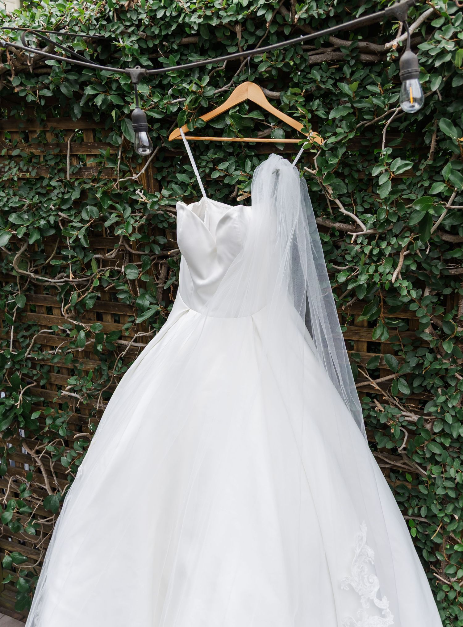 White tulle wedding dress hanging on wooden hangers against an ivy-covered wall.