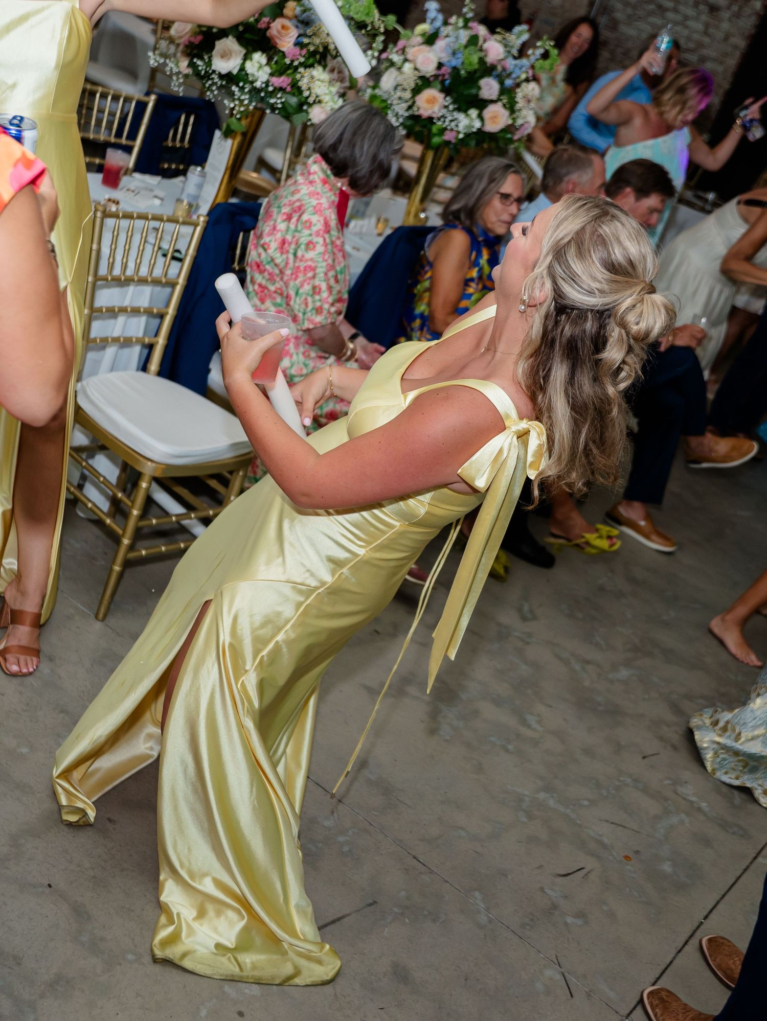 Wedding guests in yellow satin dresses participate in a fun reception game or dance activity.