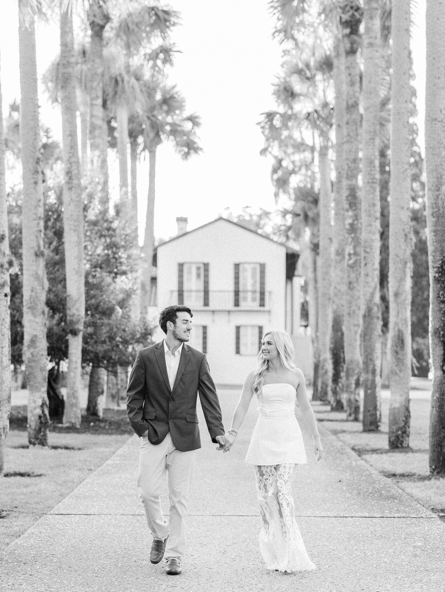 A couple walking hand in hand down a palm tree-lined path in front of a white house.