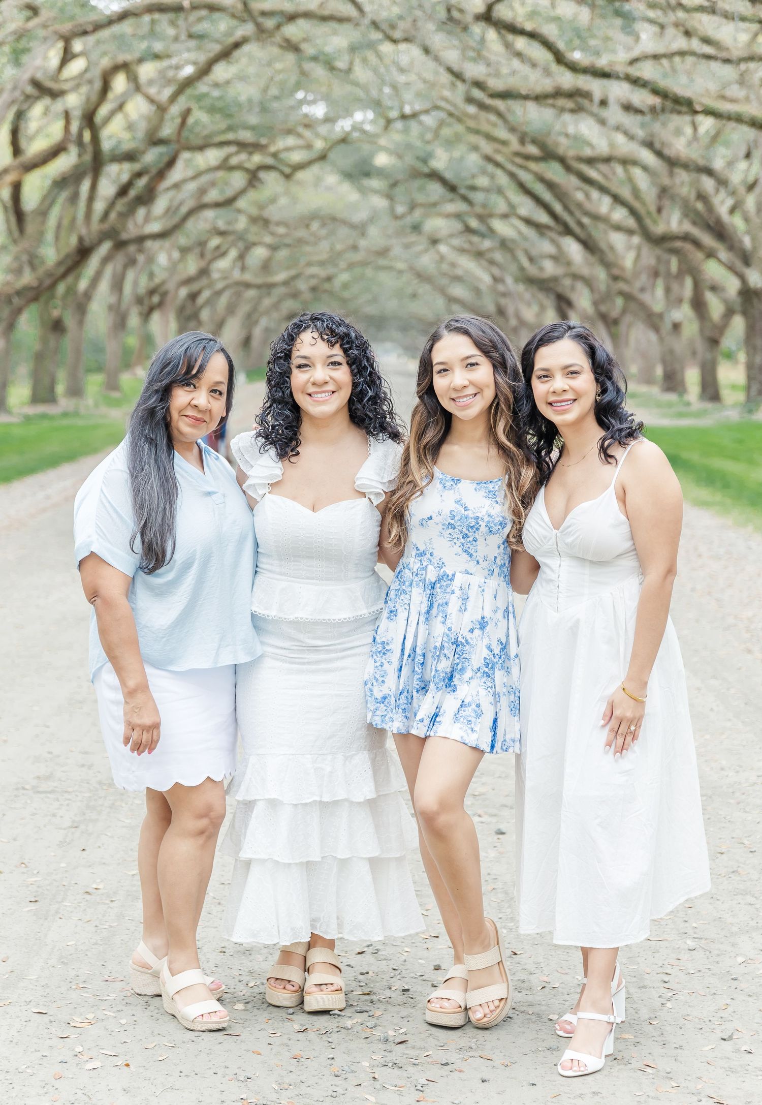 Four women in white and blue dresses pose together under an oak tree canopy.