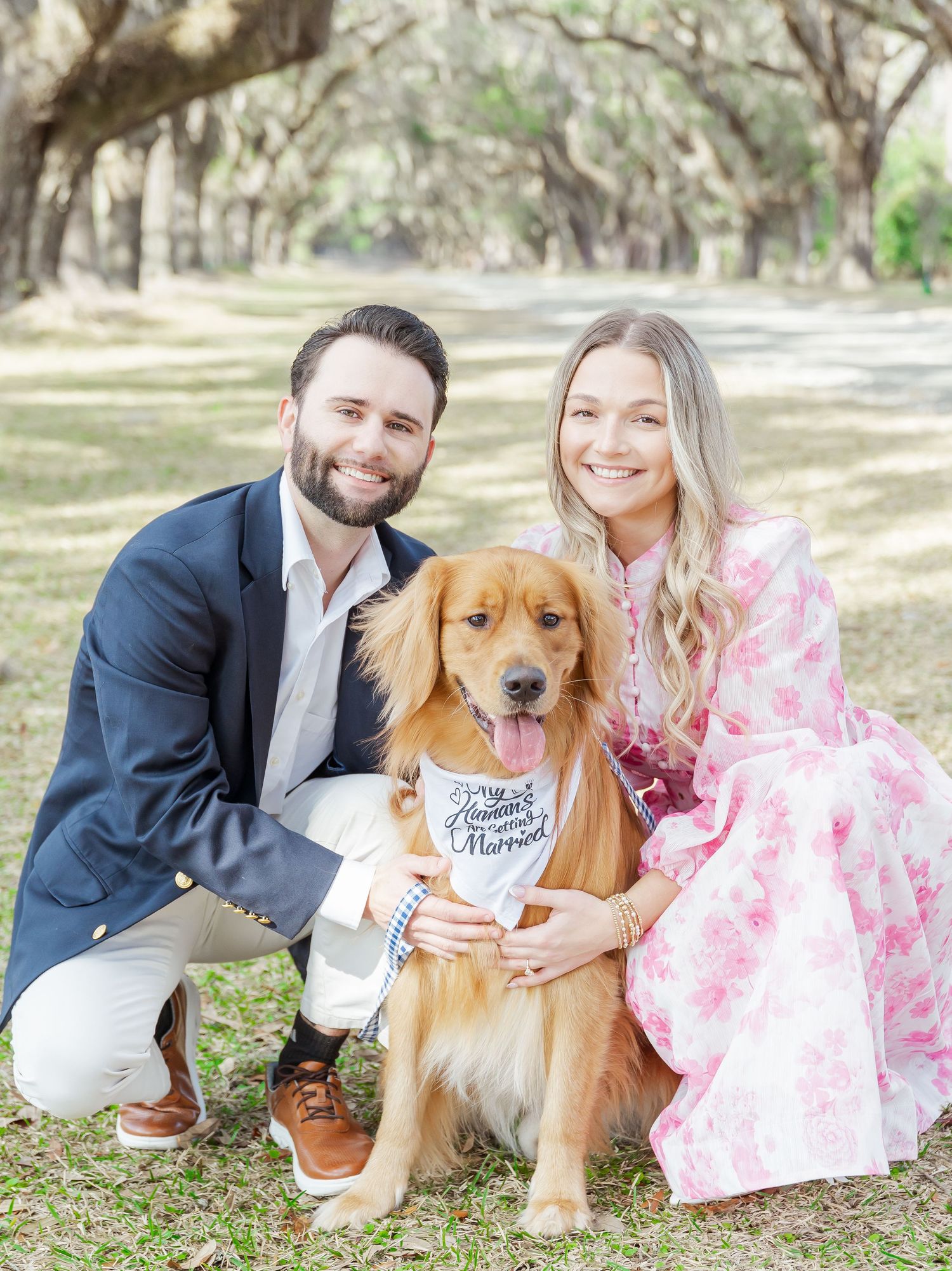 A couple poses outdoors with their golden retriever dog holding a pregnancy announcement sign under oak trees.