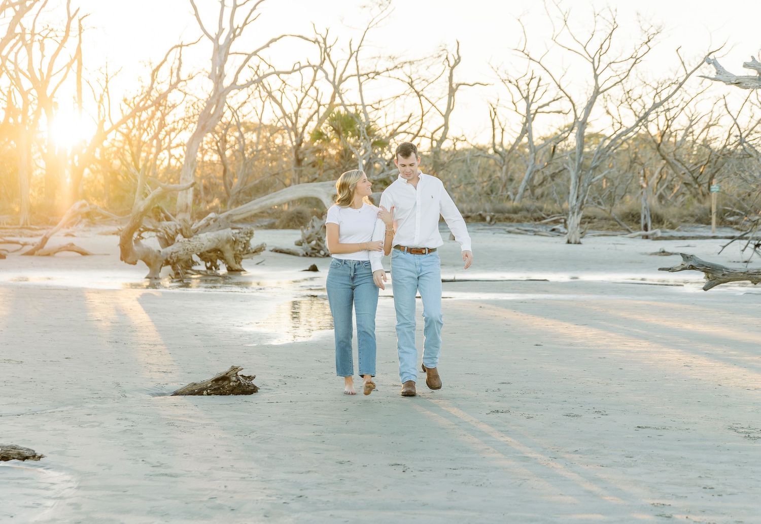 A couple strolls hand in hand on a beach with driftwood and sunset in background.