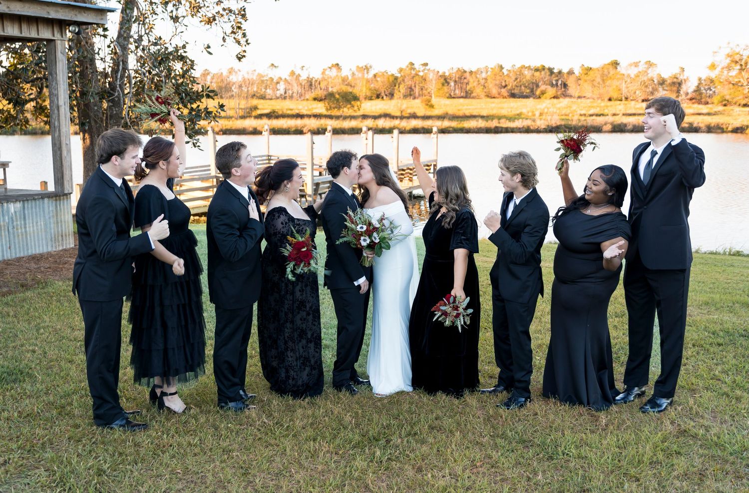 Wedding party laughs together during group photos by the lake at sunset.