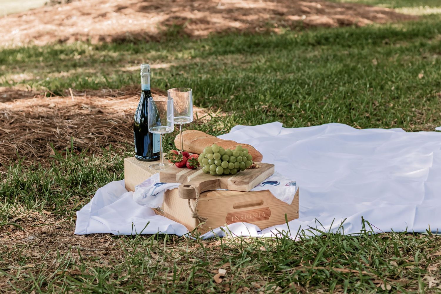 A picnic spread on a white blanket with wine, drinks and food in a grassy outdoor setting.