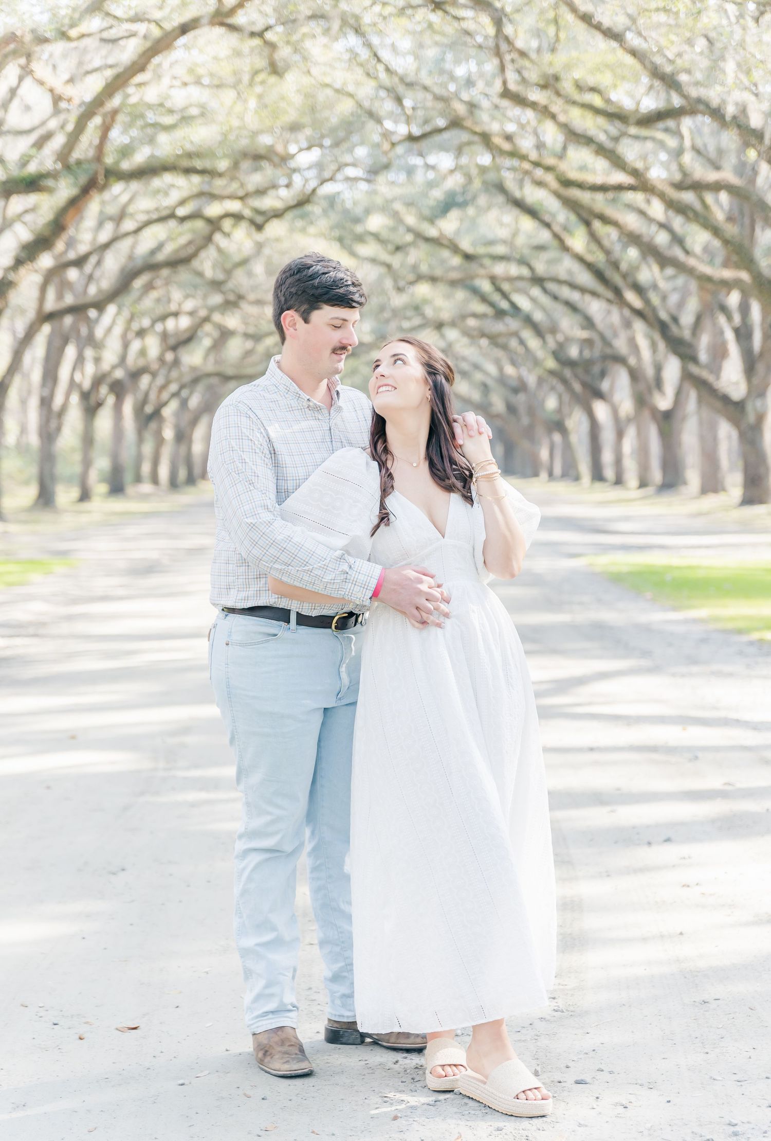 A couple embraces on a scenic oak-lined path wearing white and light blue casual attire.