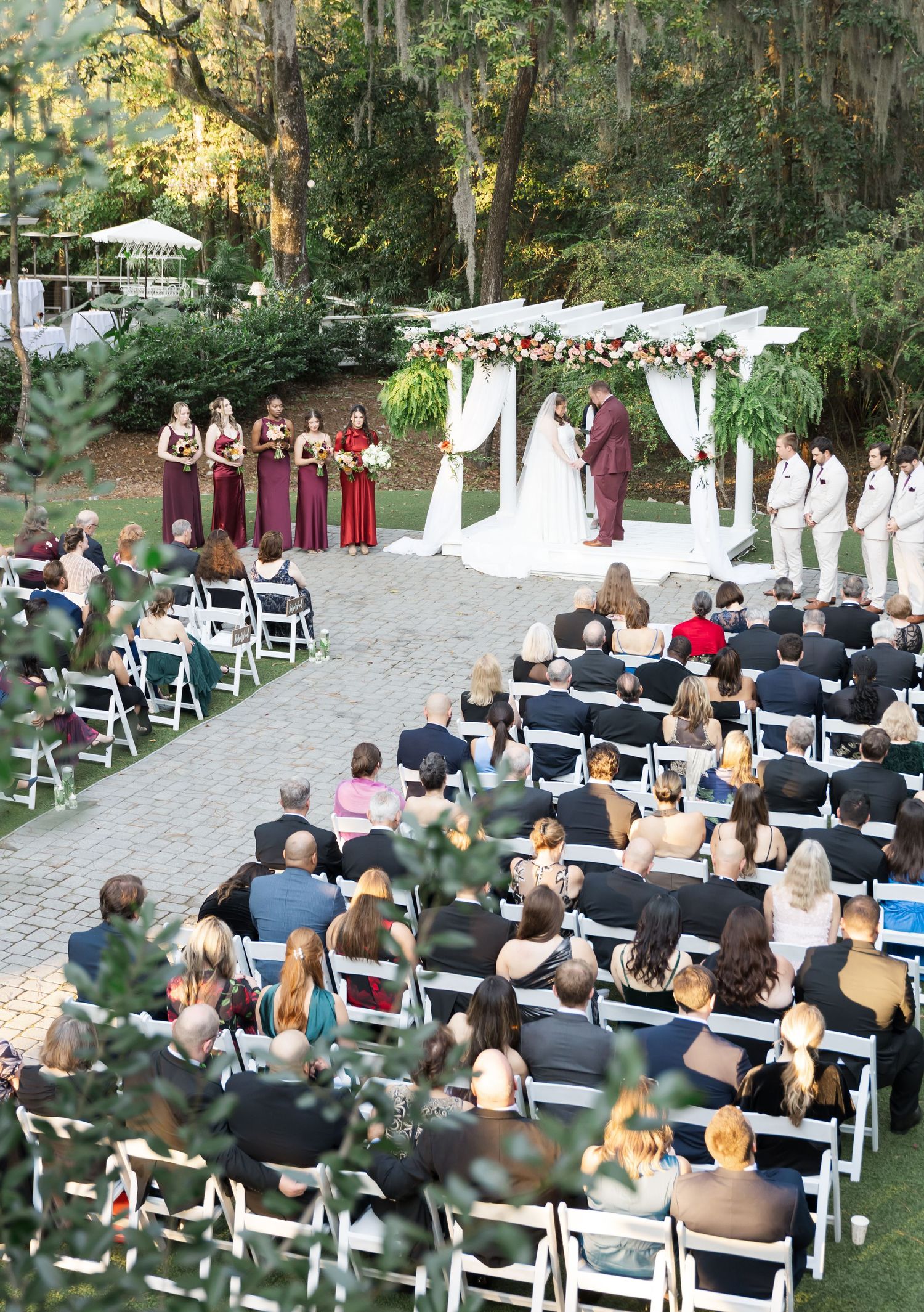 Outdoor wedding ceremony setup with white chairs and floral decorations under a canopy of trees.