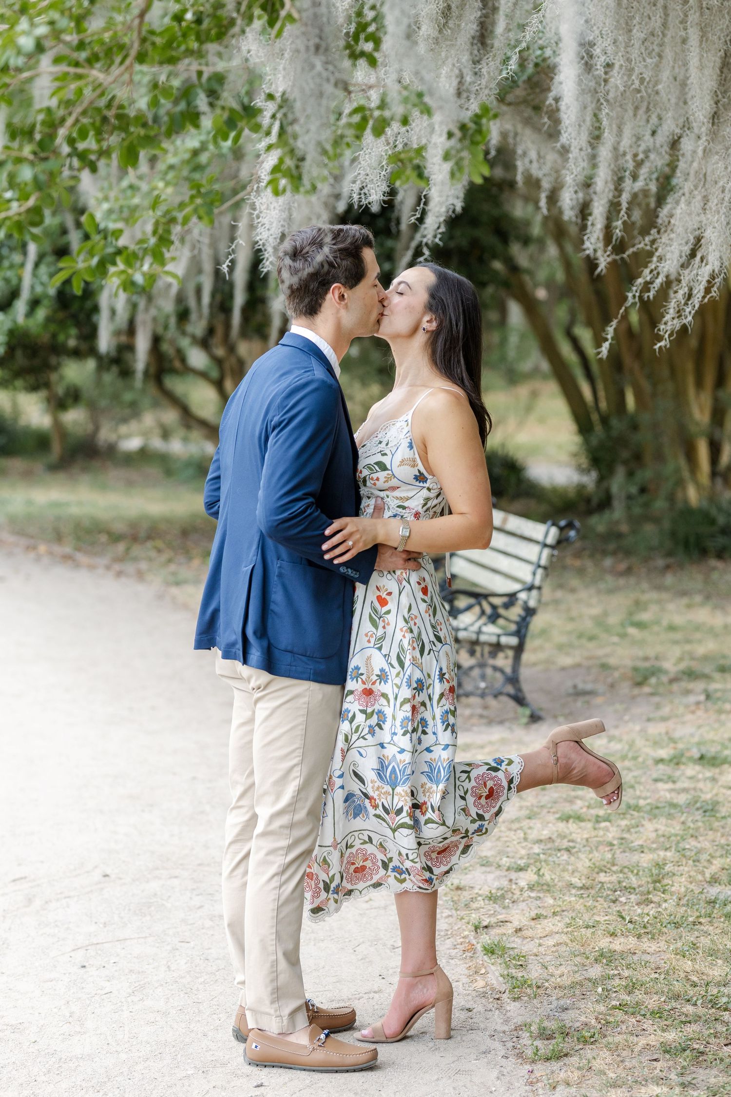 Couple sharing a kiss under Spanish moss-draped trees on a garden path, woman in floral dress, man in navy blazer.