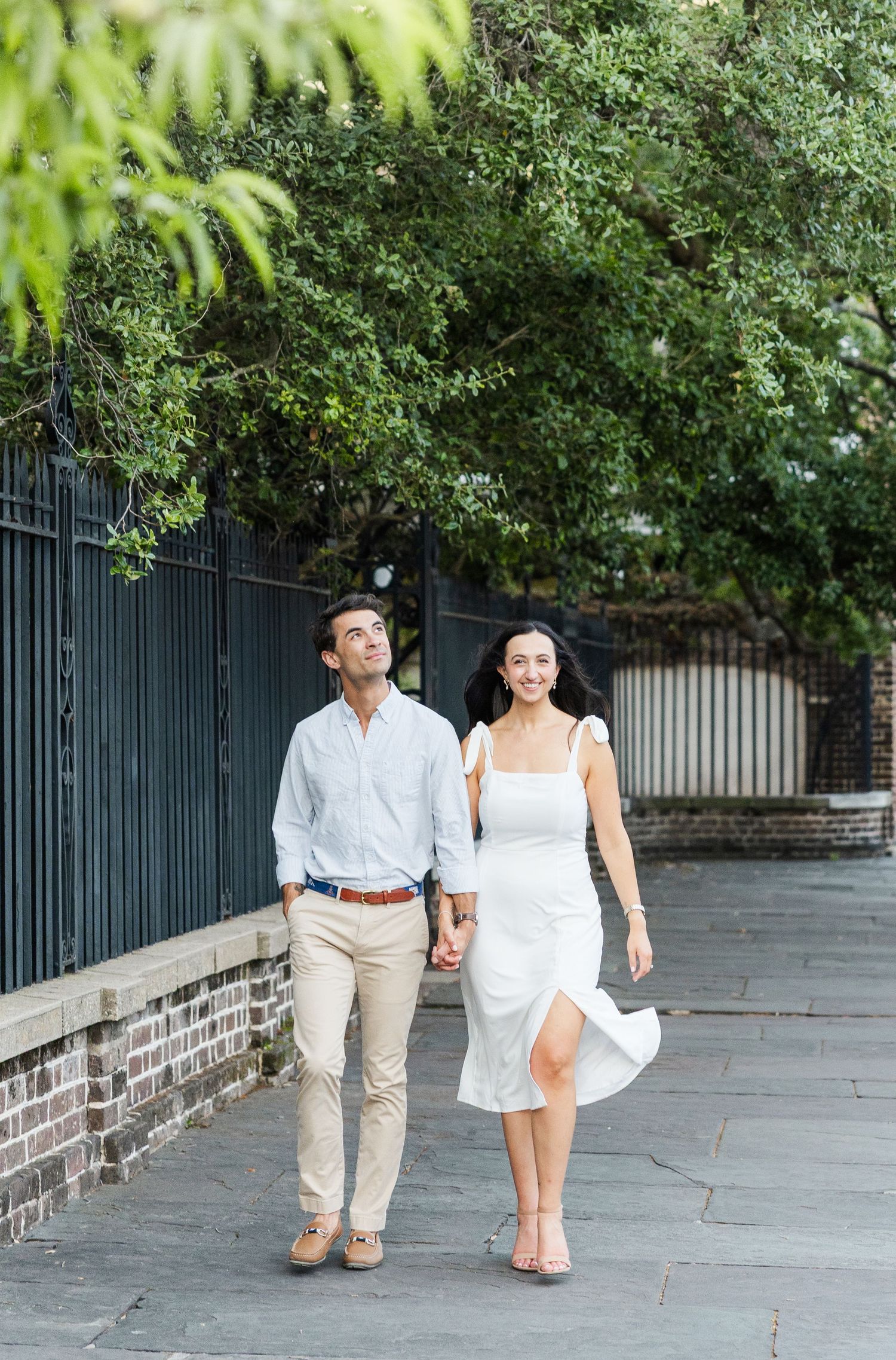 Happy couple walking hand in hand on a historic cobblestone sidewalk beside an iron fence and trees.