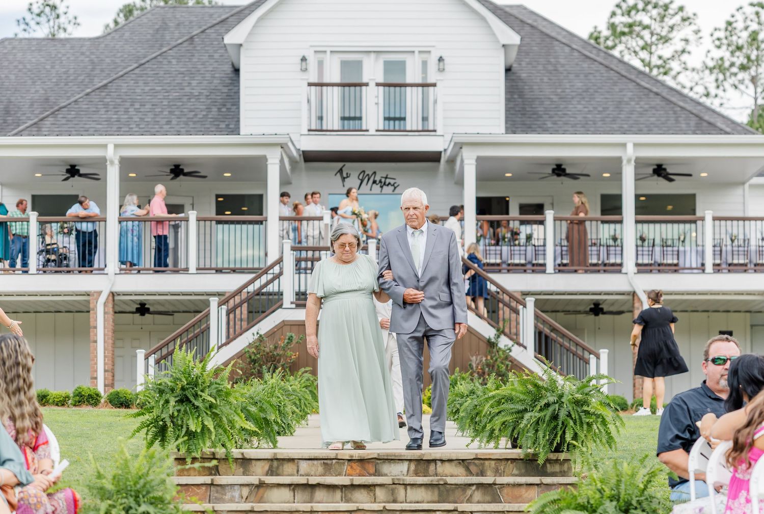 A couple walks down stone steps at a white farmhouse wedding venue during an outdoor ceremony.