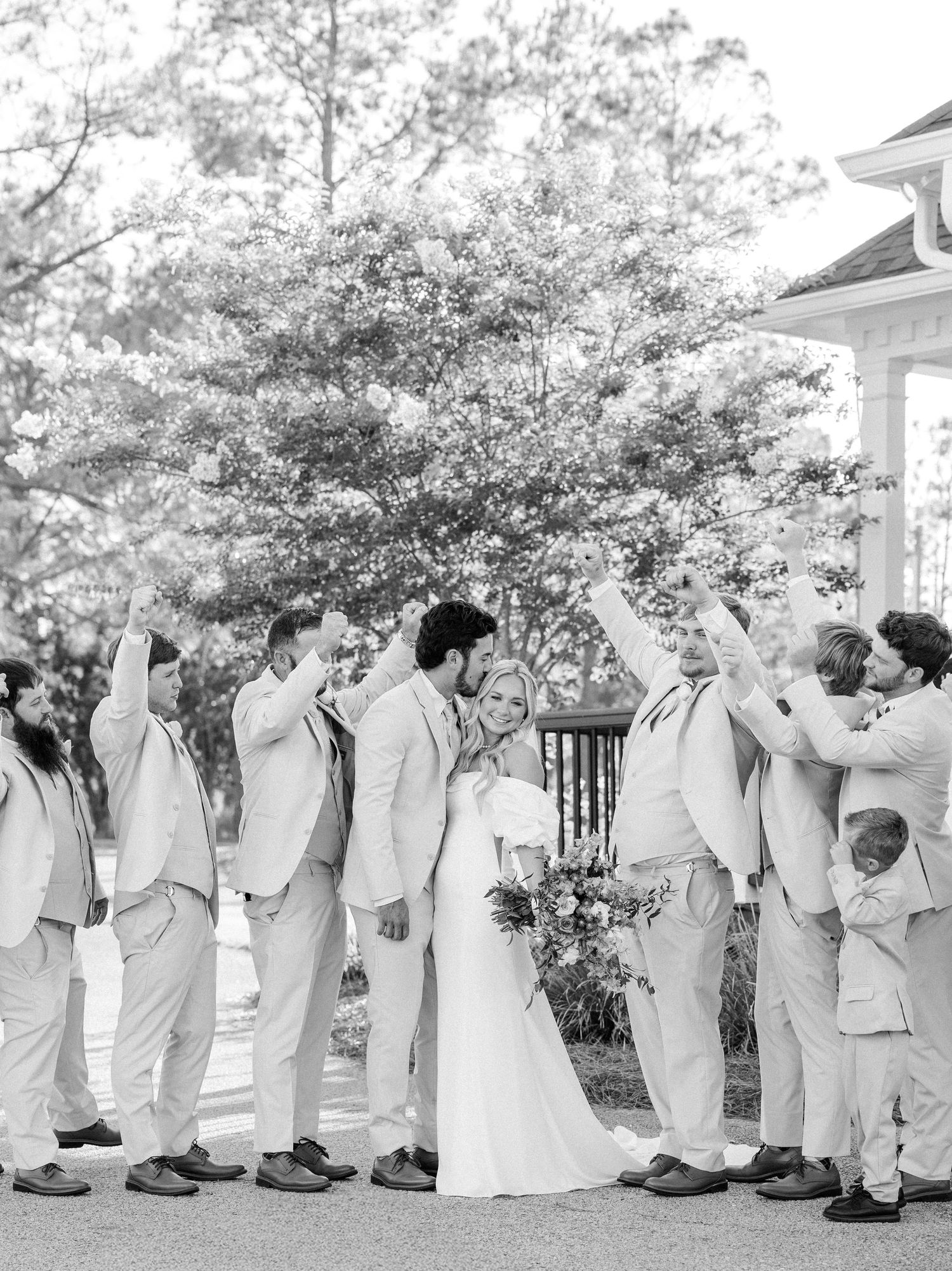 Black and white photo of bride and groom celebrating with groomsmen in matching light suits outdoors.