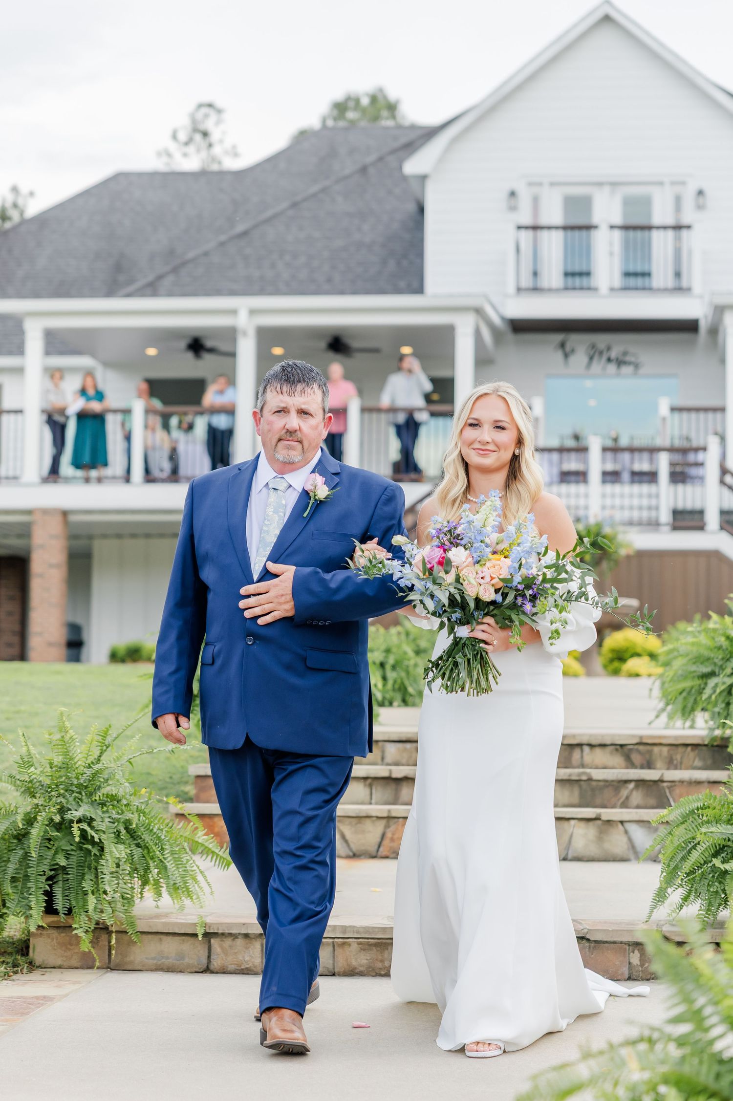 Bride in white gown holding colorful bouquet walks with man in navy suit outside elegant venue.