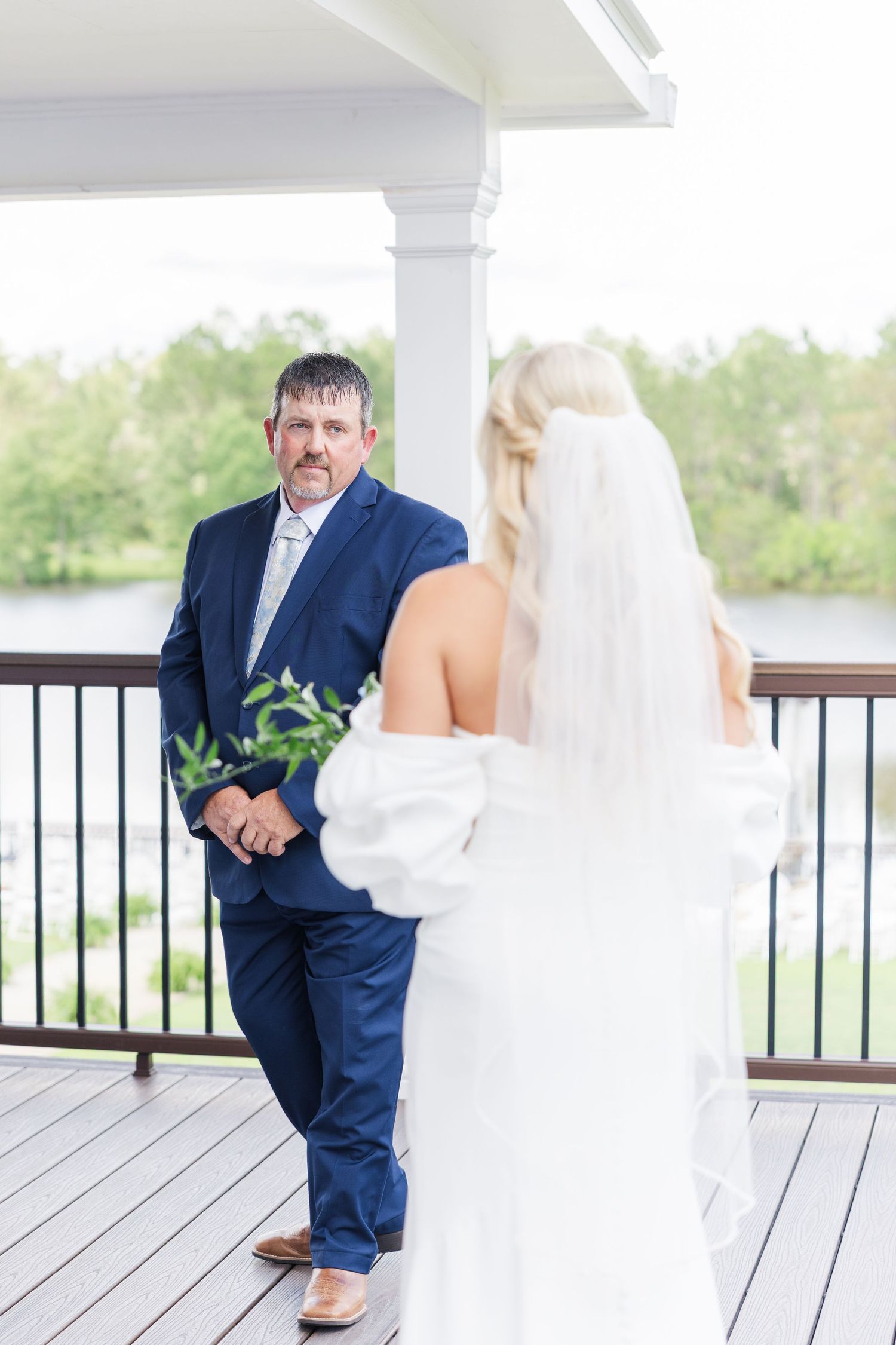 A bride in a white gown and veil shares an emotional first look moment with a man in a navy suit on a deck.