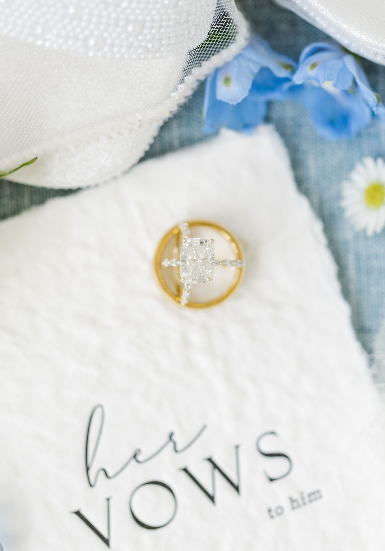 Gold wedding band and diamond engagement ring resting on a white vows book with blue flowers and lace in the background.