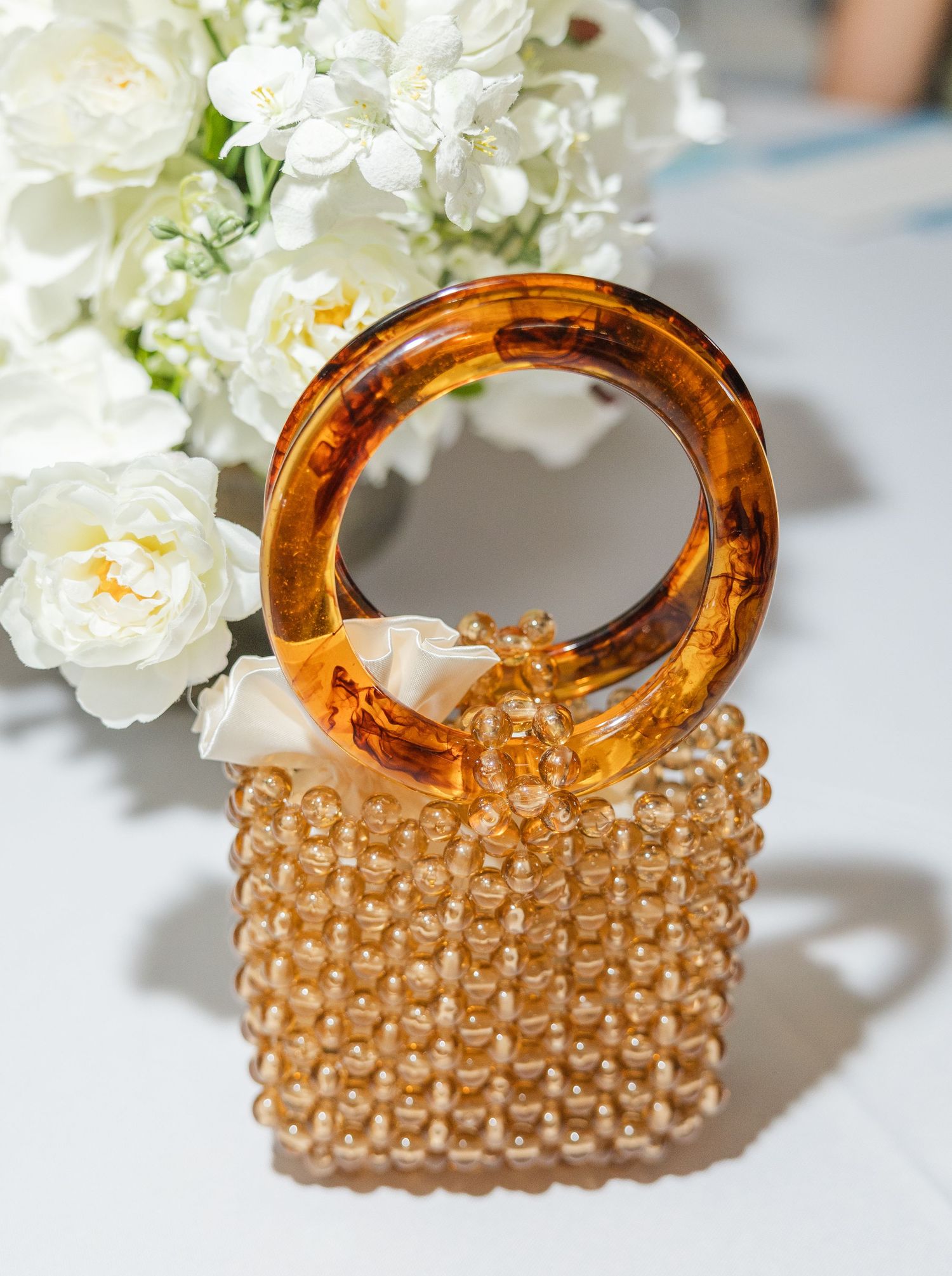 Gold beaded mini bag with tortoiseshell acrylic ring handle and ivory bow, styled with white flowers.