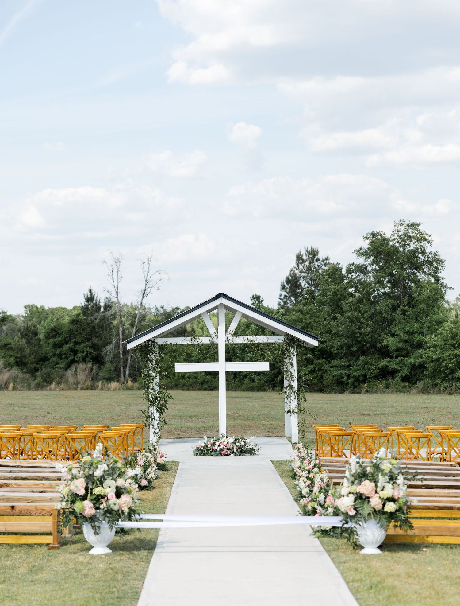 Outdoor wedding ceremony setup with white cross altar, floral arrangements, and wooden benches on green lawn.