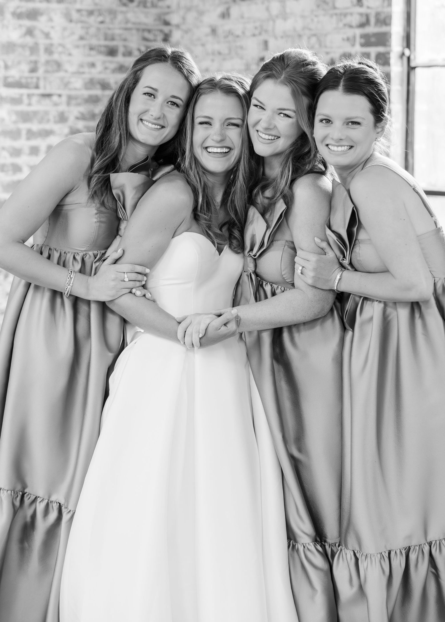 Black and white photo of bride in white gown embraced by three bridesmaids in satin dresses.