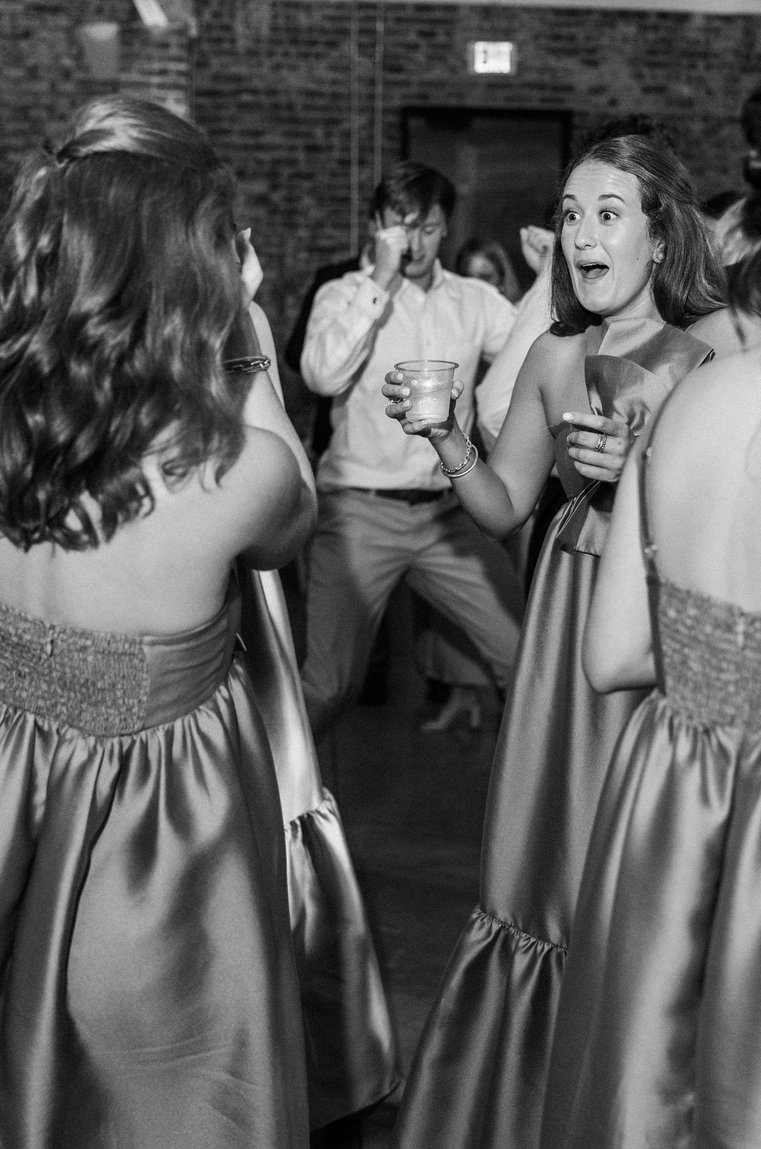 Two bridesmaids in satin gowns dancing and laughing at wedding reception in black and white photo.