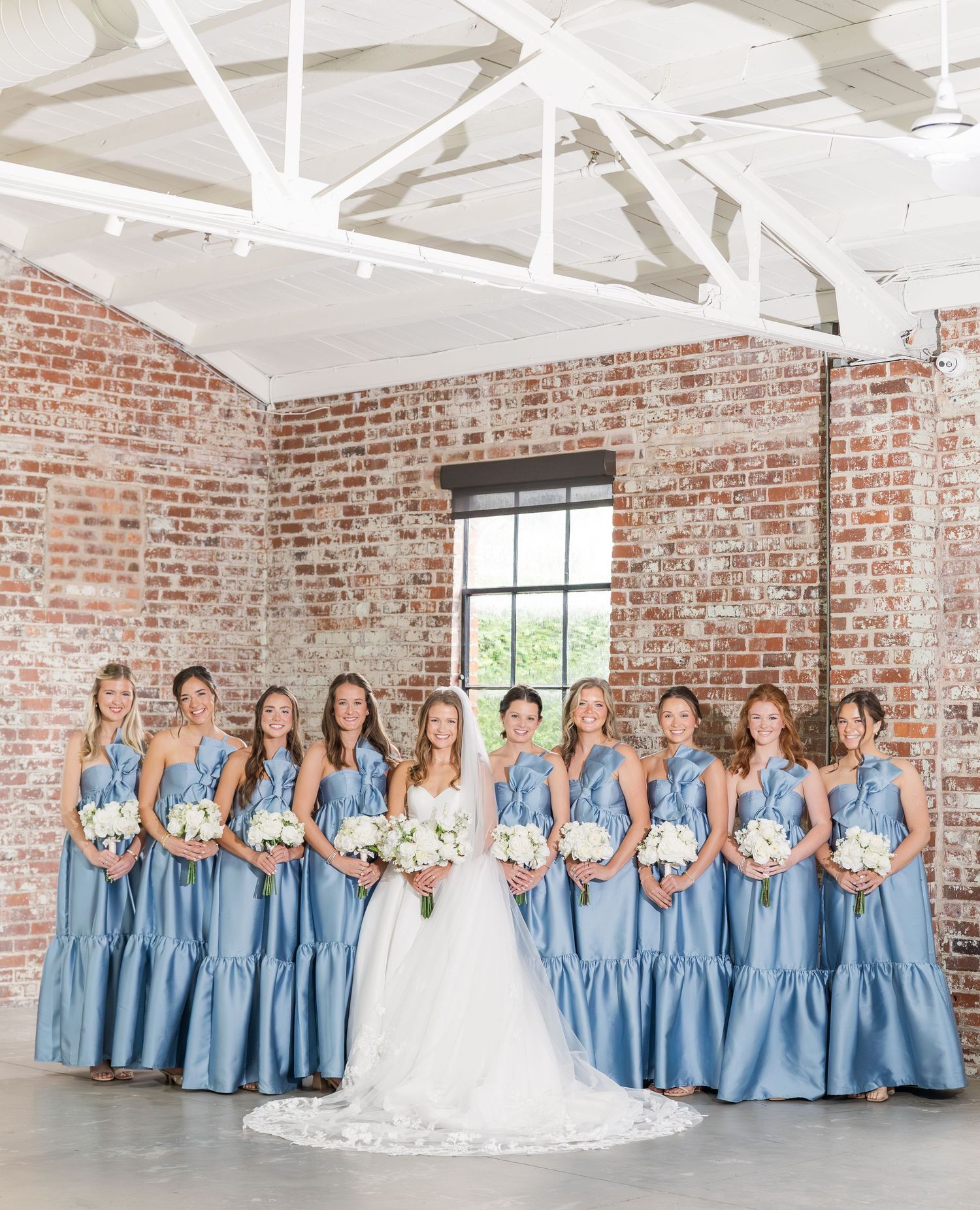 Bride in white gown poses with ten bridesmaids in dusty blue tiered satin dresses in brick venue.
