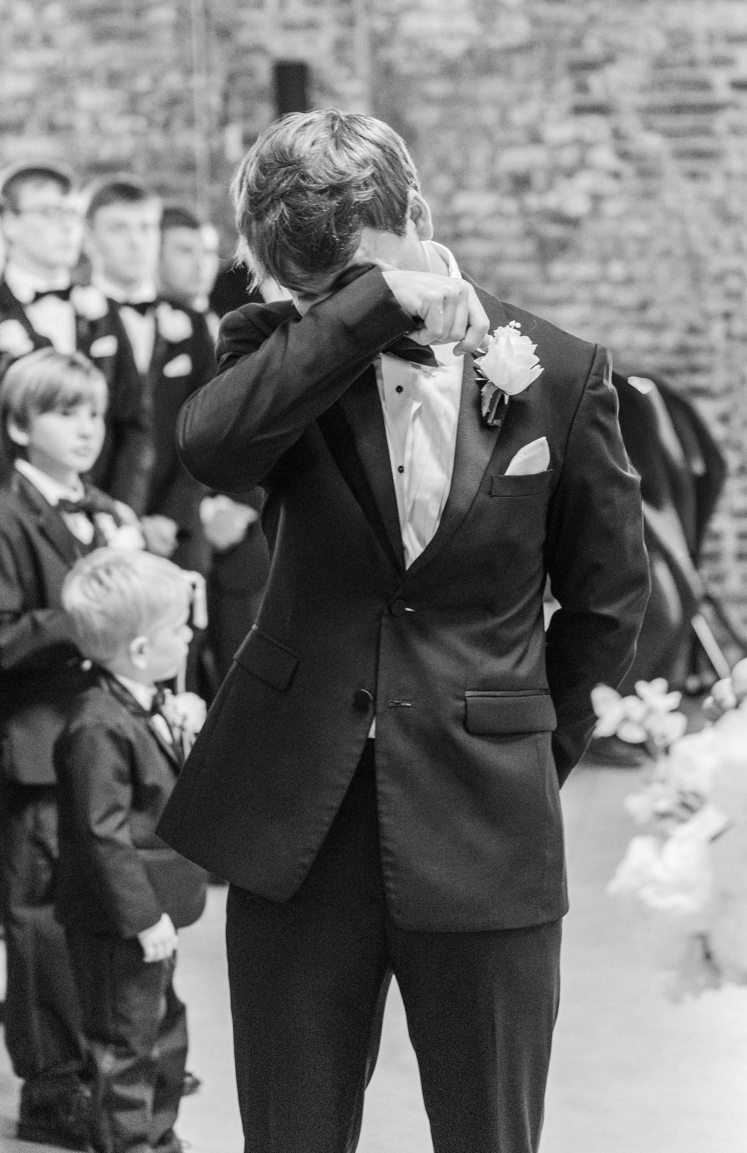 Emotional groom in black tuxedo wipes tears at altar surrounded by groomsmen and ring bearers.