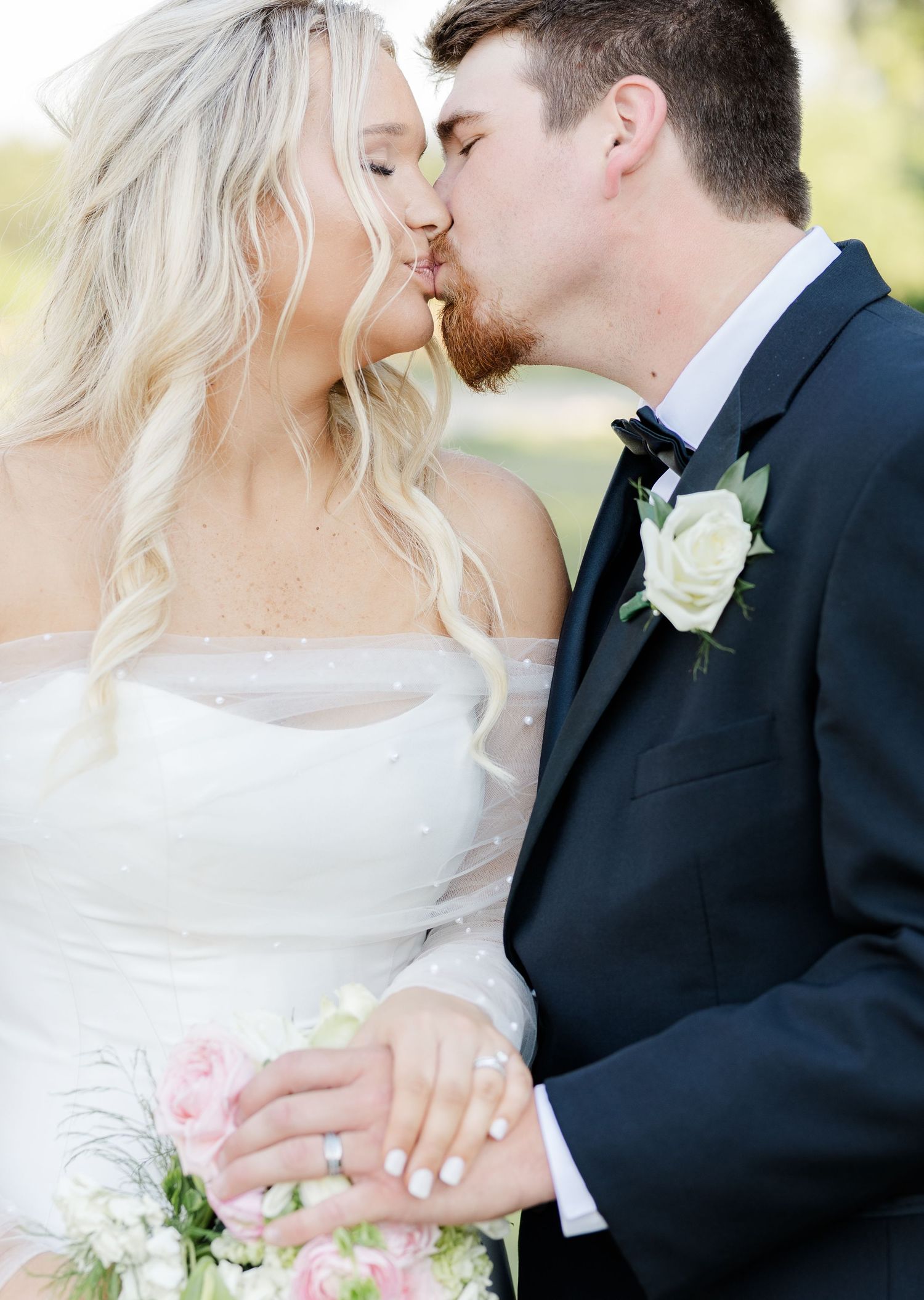 Bride and groom sharing romantic kiss outdoors, bride holding white and pink floral bouquet in wedding attire.