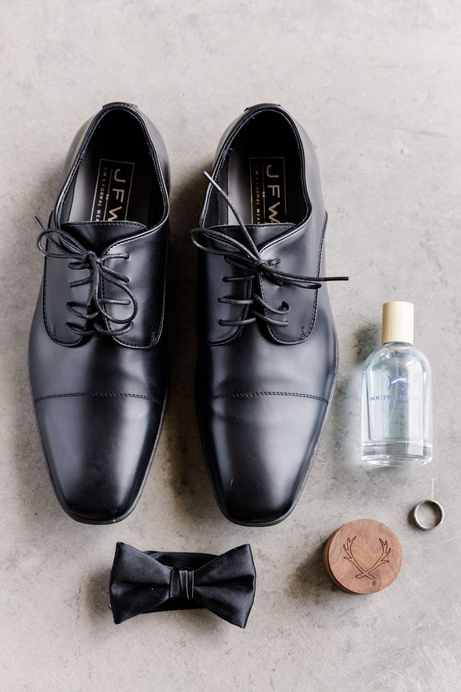 Groom wedding details flat lay with black oxford shoes, bow tie, cologne, and gold wedding band.