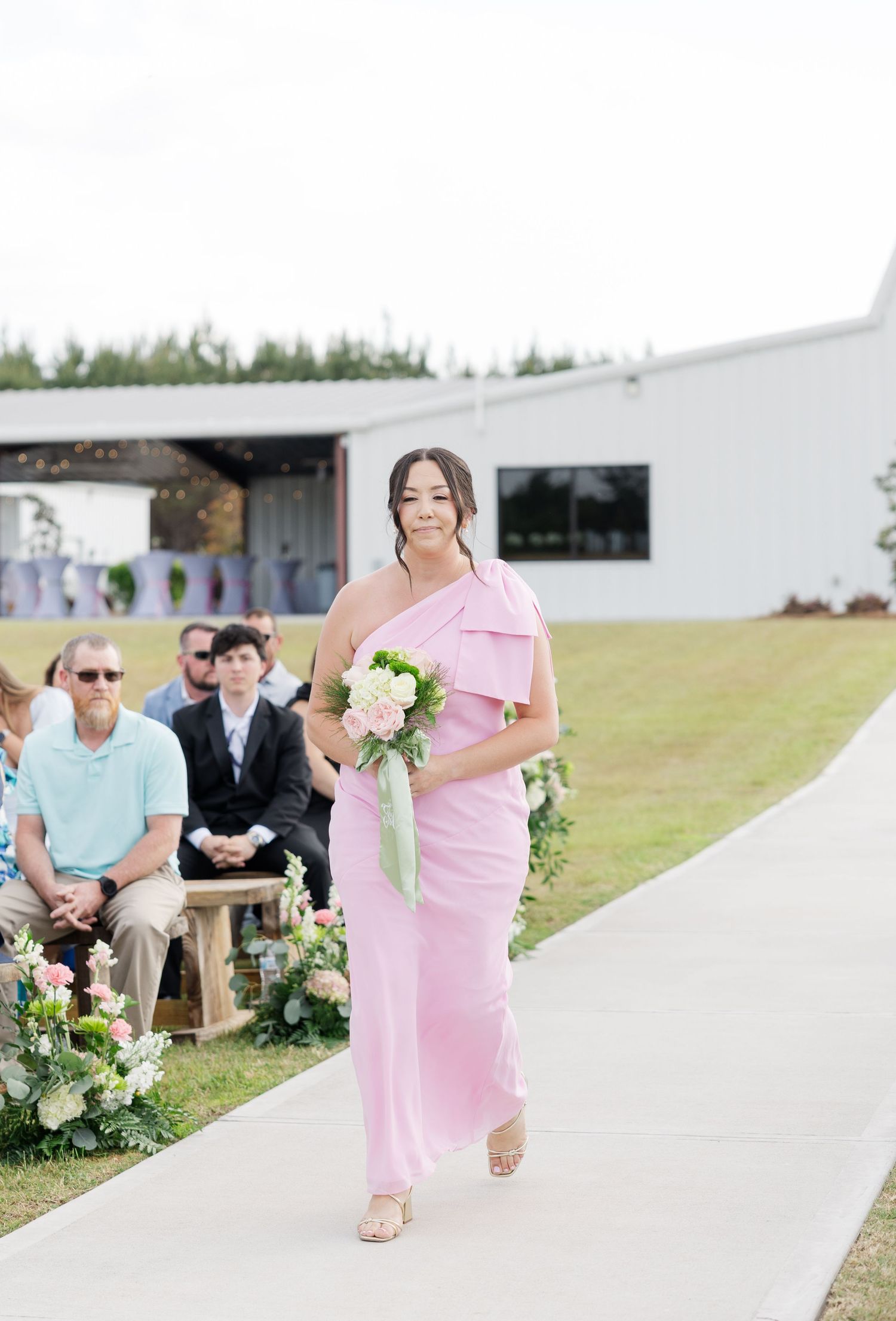 Bridesmaid in pink one-shoulder dress walking down outdoor wedding aisle with bouquet.