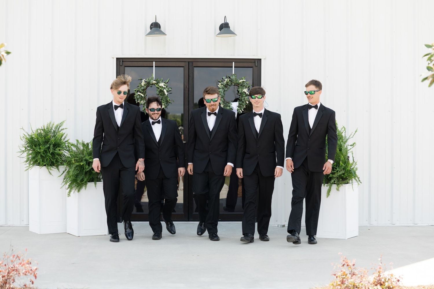 Five groomsmen in black tuxedos and bow ties standing before white barn doors with greenery.