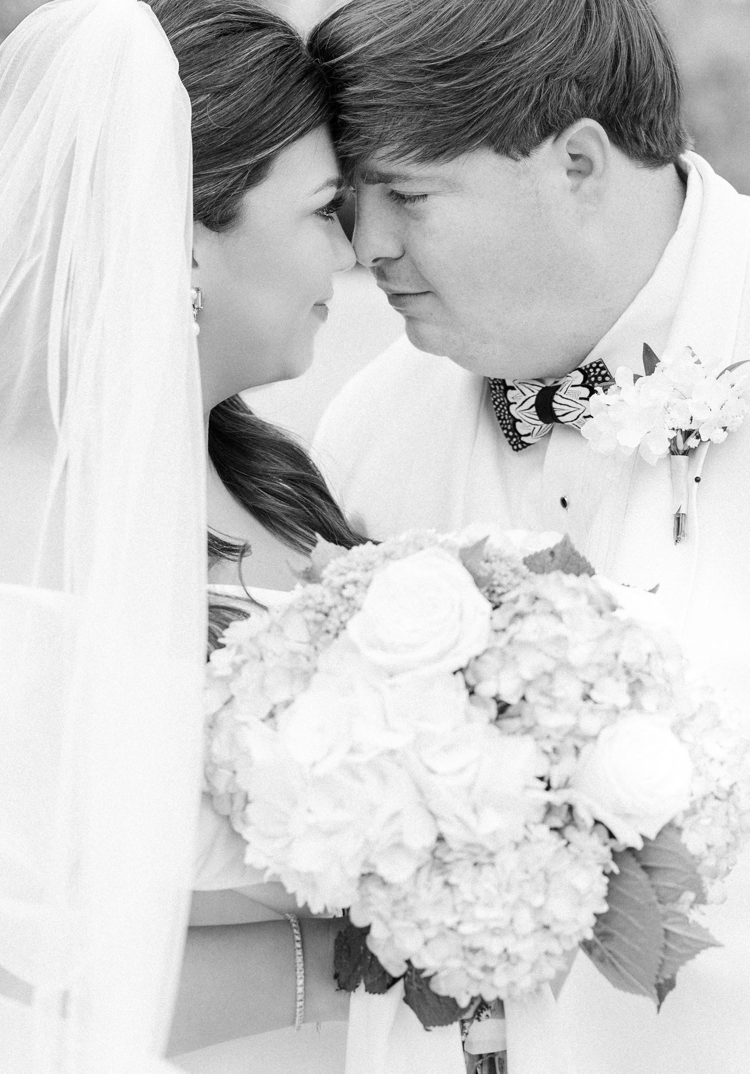 Black and white romantic portrait of bride and groom touching foreheads with white rose bouquet.