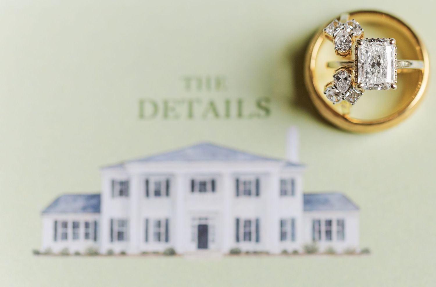 Diamond engagement ring and wedding band displayed on gold dish atop elegant wedding details card with venue illustration.