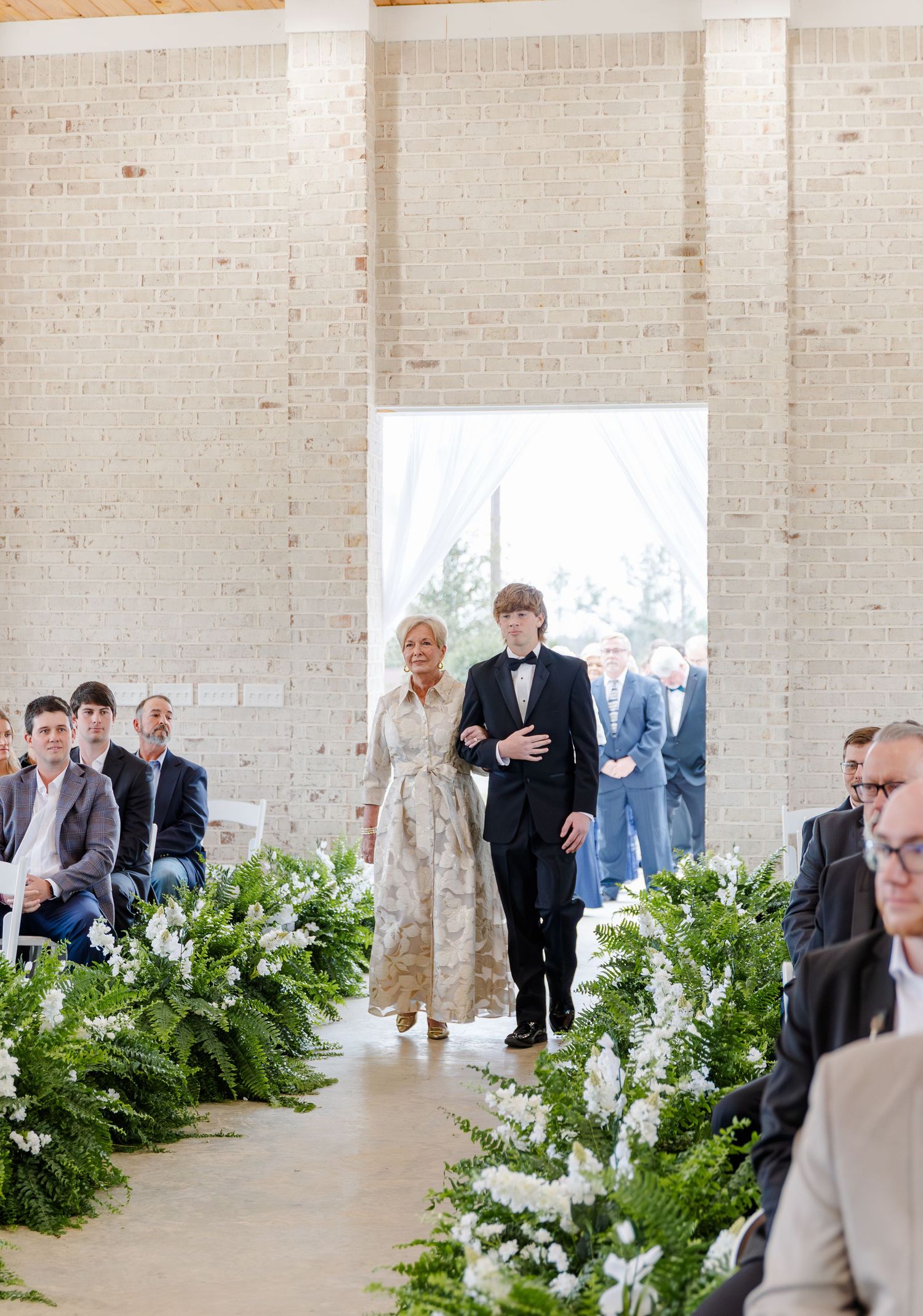 Wedding guests walk down aisle decorated with lush greenery and white flowers in rustic brick ceremony venue.