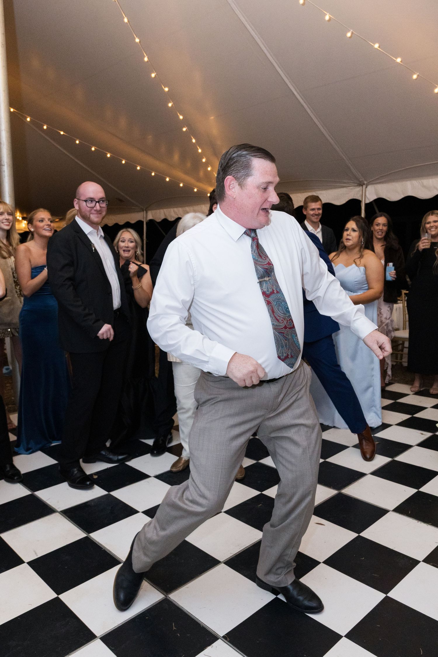 Man energetically dancing on black and white checkered floor at wedding reception under a tent with string lights.