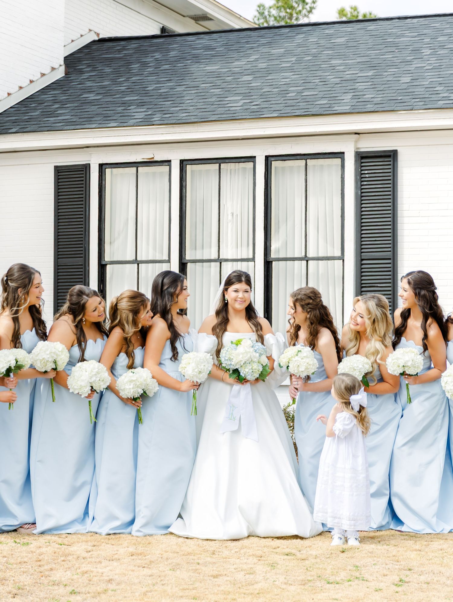 Bride in white gown surrounded by bridesmaids in light blue dresses holding white hydrangea bouquets.
