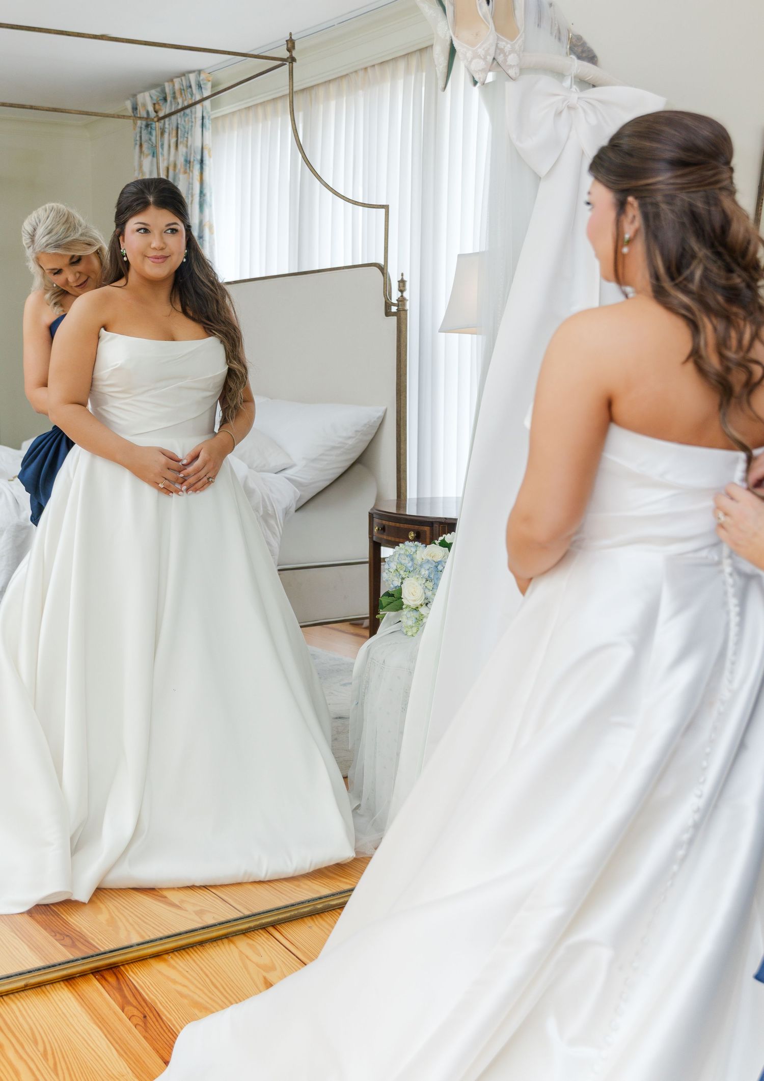 Bride in strapless white ball gown admiring her reflection in mirror with bridesmaid assisting on wedding day.