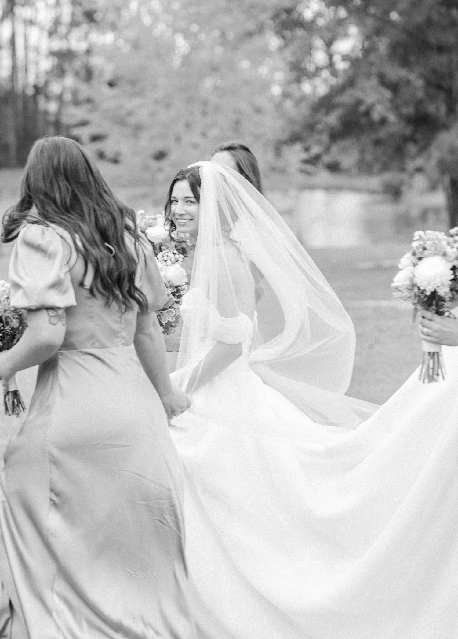 Black and white photo of bridesmaids holding up the bride's flowing veil in an outdoor setting.