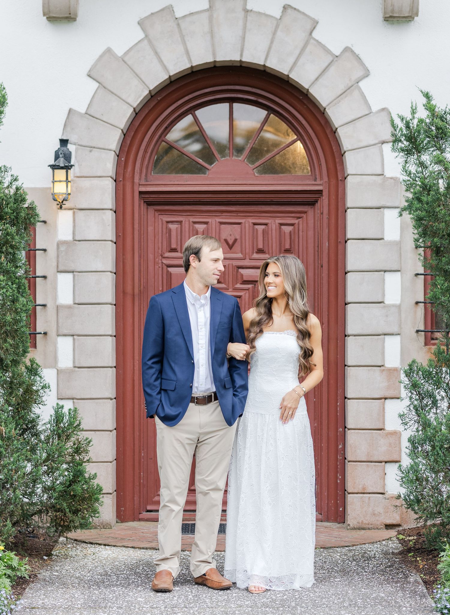Couple in elegant attire poses before ornate arched red door with stone trim and lush greenery.