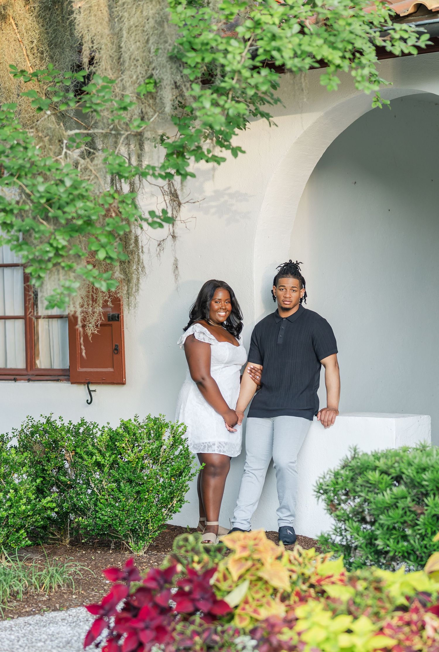 A couple poses together in front of a white arched building surrounded by lush greenery and colorful flowers.