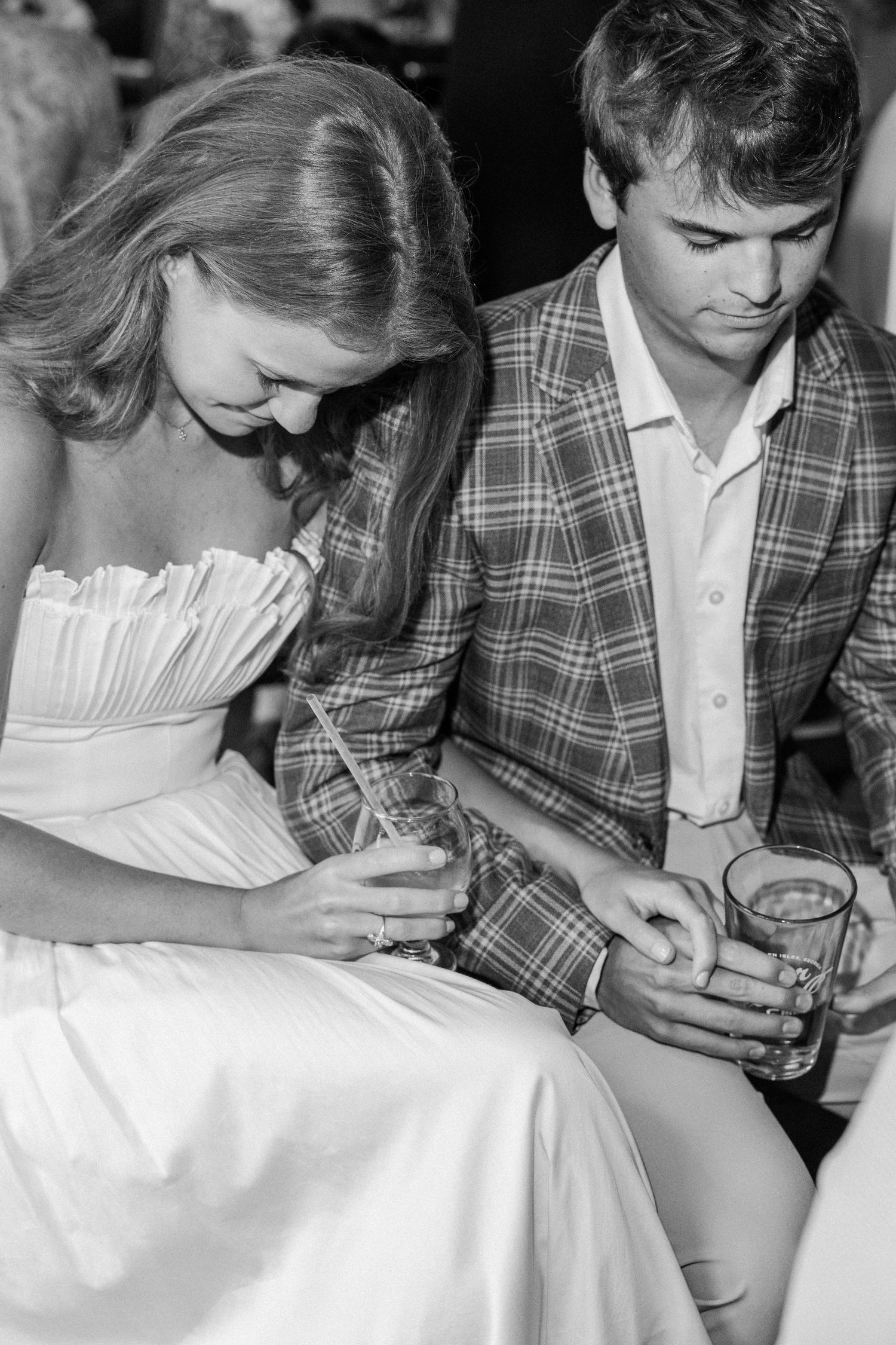 Black and white photo of a bride and groom sitting together holding drinks at their wedding reception.