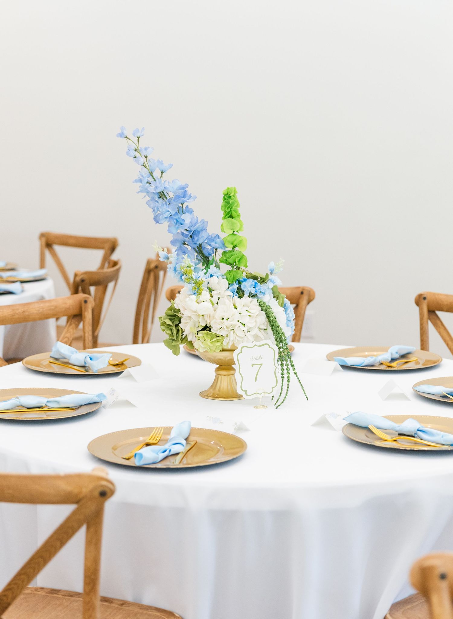 Elegant wedding table with white linen, gold chargers, blue napkins, and a floral centerpiece of blue and white blooms.