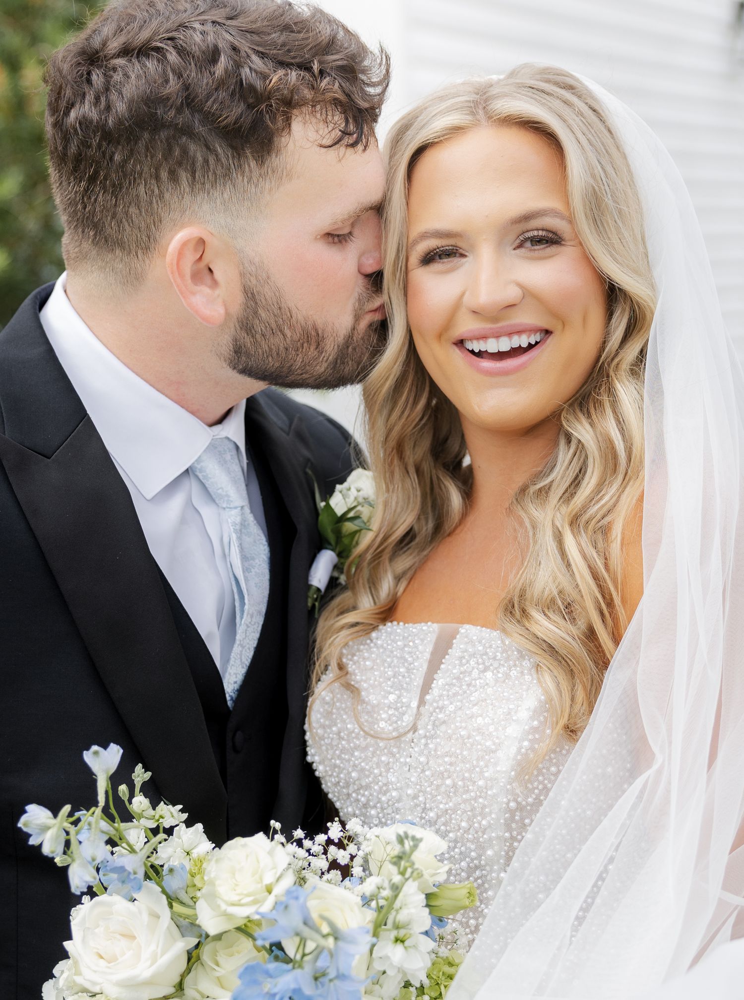 Groom kissing bride on cheek as she smiles holding white and blue floral bouquet at wedding.