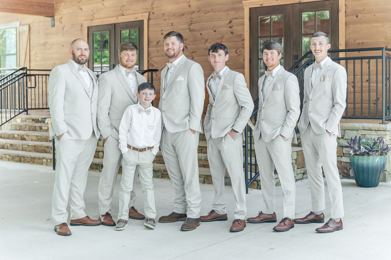 Groomsmen in light beige suits pose with young ring bearer on wooden porch.