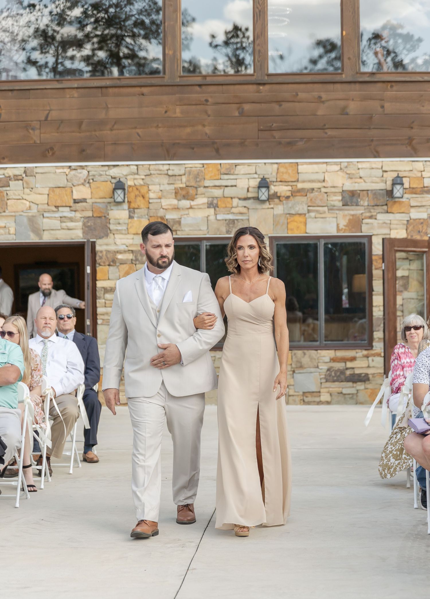 A couple walks arm in arm at an outdoor wedding ceremony wearing beige and cream formal attire.